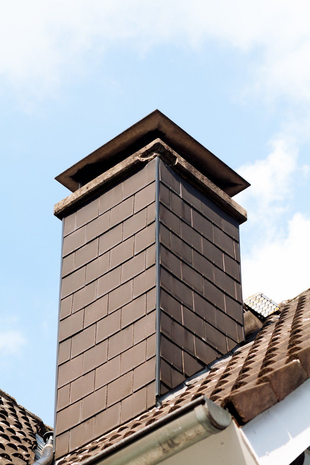 Brick chimney on a rooftop emitting white smoke against a backdrop of houses.