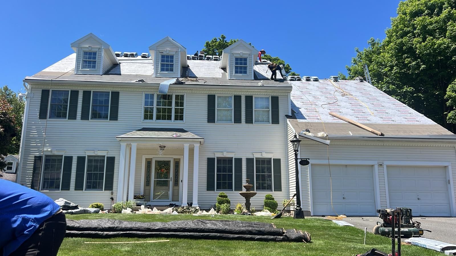 House with roof partially replaced; white exterior, green shutters, garage, blue sky.