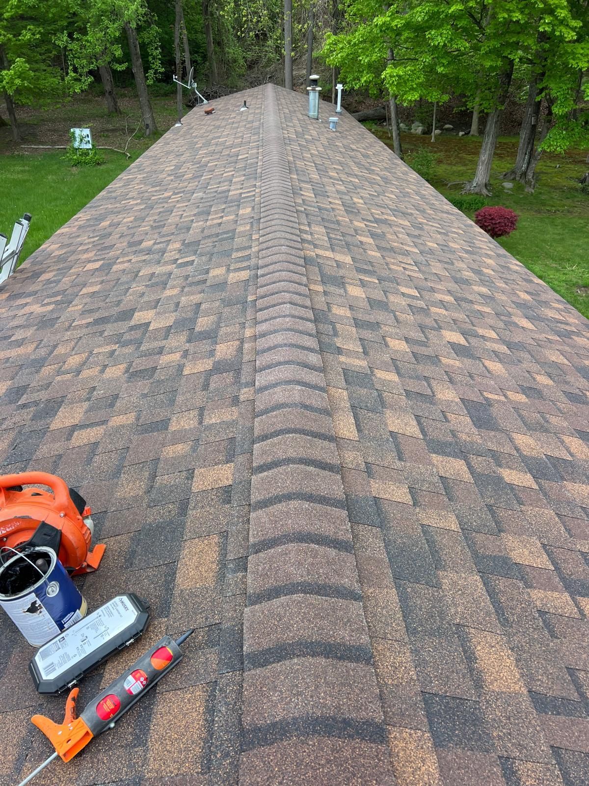 Brown shingle roof with orange and black tools. Green trees and grass in the background.