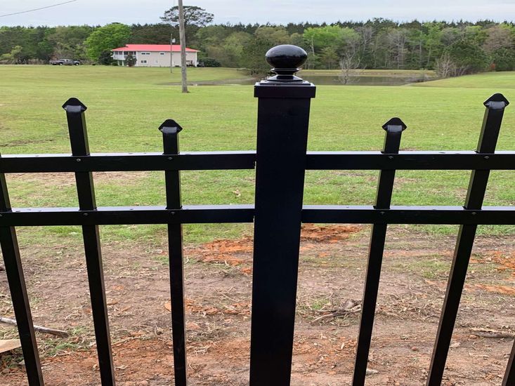 Black metal fence with a grassy field and distant house under an overcast sky