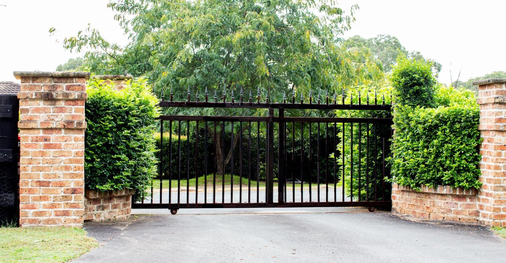 Black metal driveway gate between brick pillars and green hedges, with trees behind.