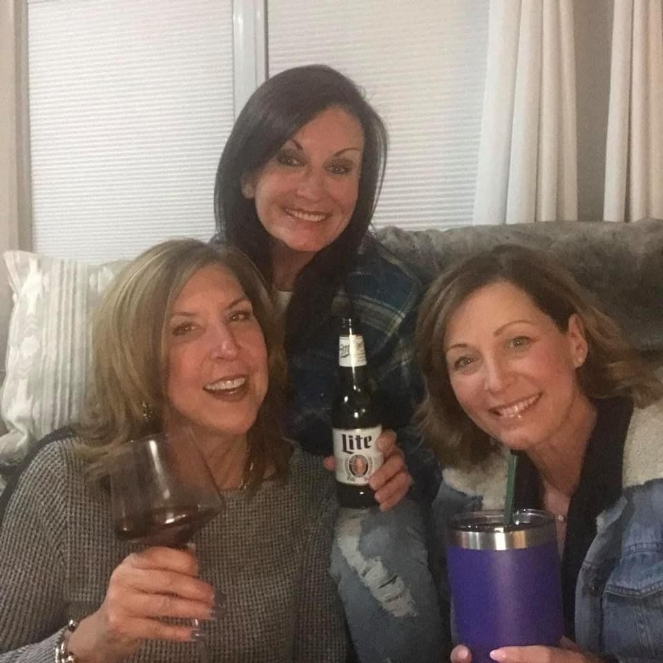 Three women are sitting on a couch holding glasses of wine and a bottle of lite beer.