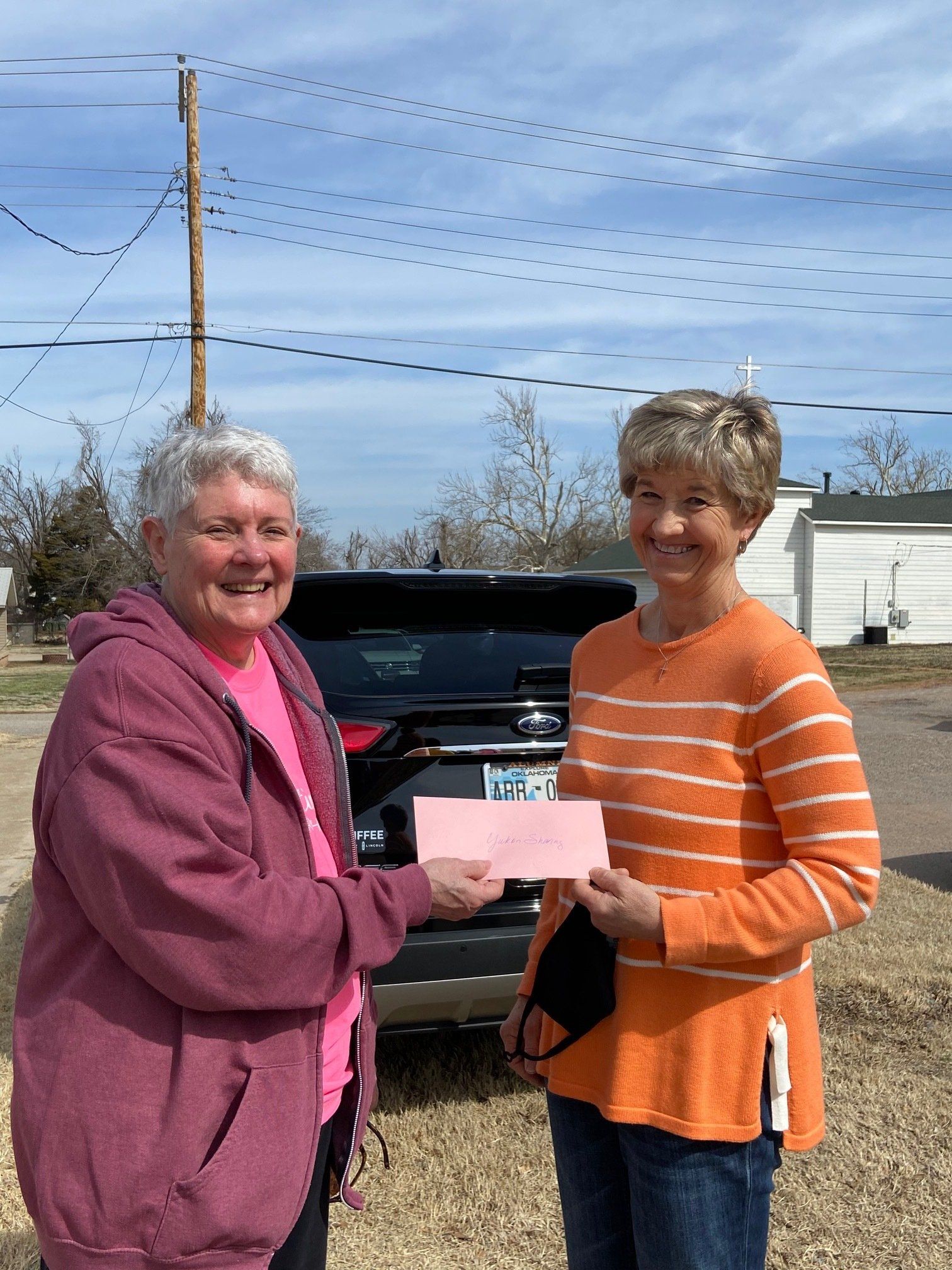 Two women are standing next to each other in front of a car holding a check.