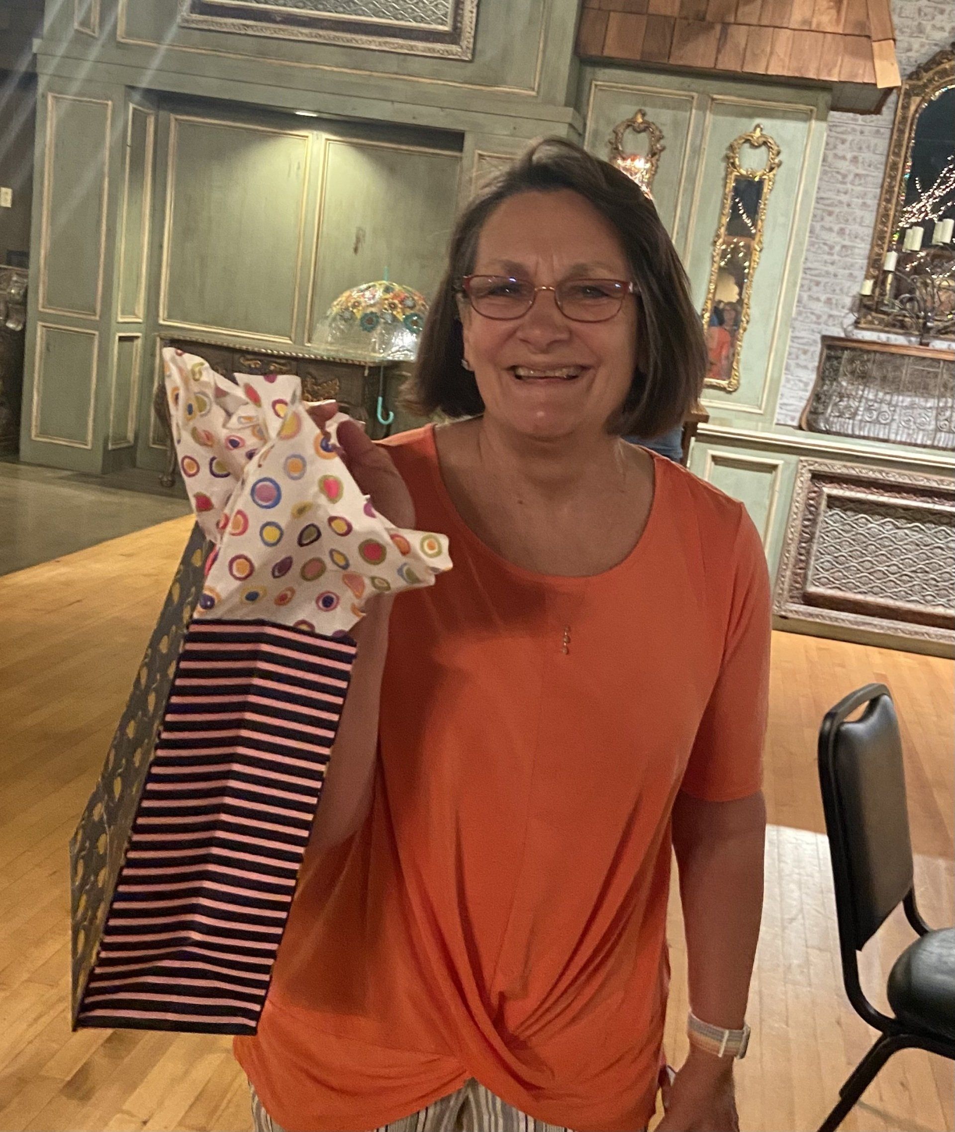 A woman in an orange shirt is holding a gift bag and smiling.