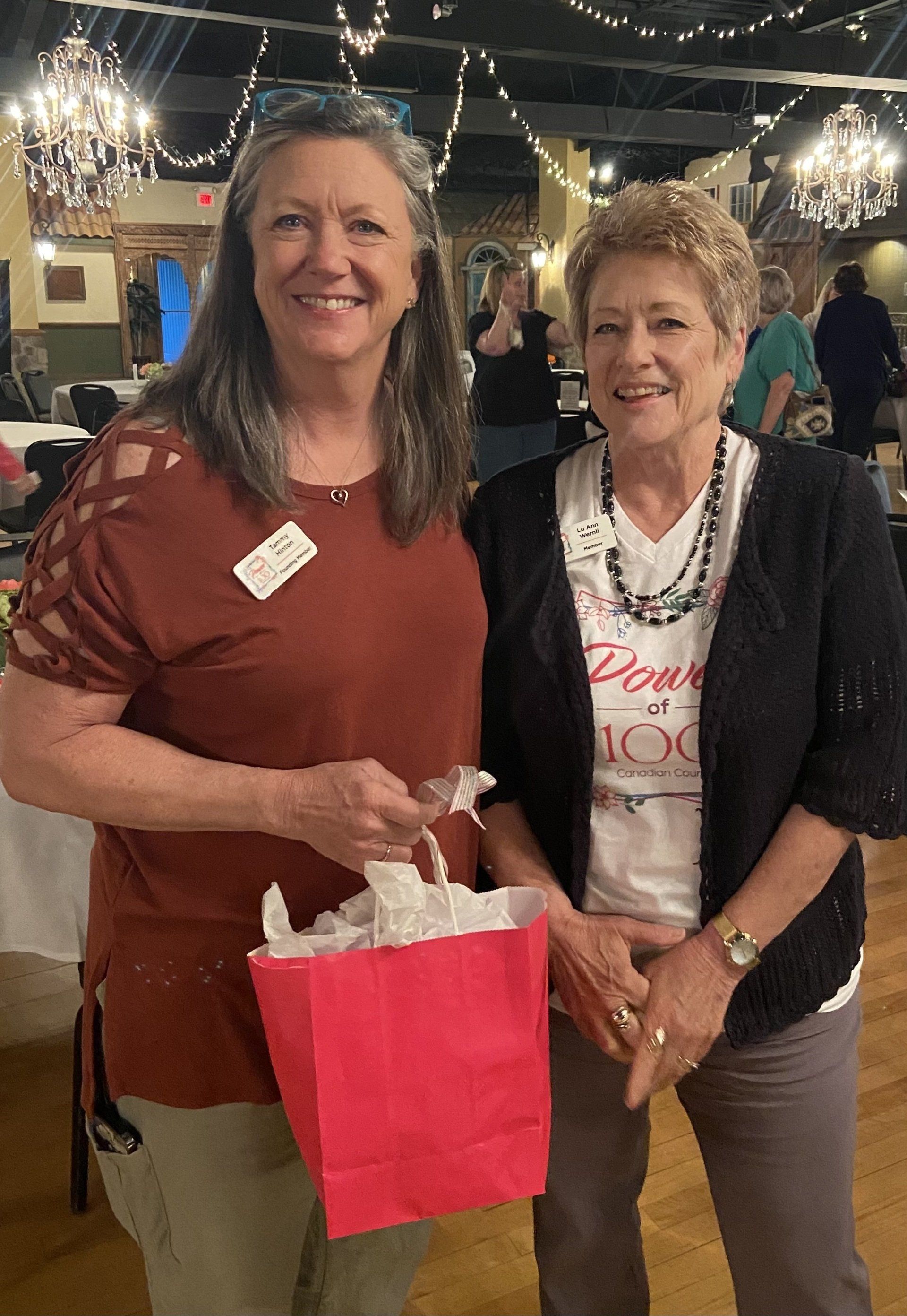 Two women are standing next to each other in a room holding a pink bag.