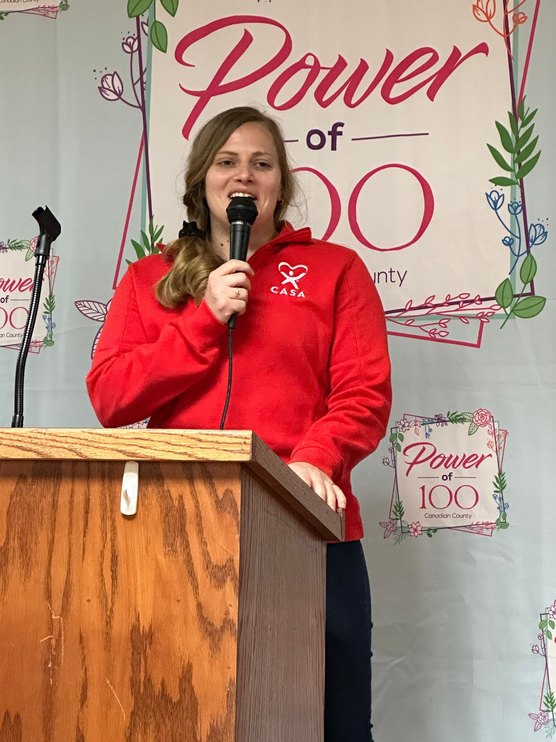 A woman in a red sweatshirt stands at a podium holding a microphone