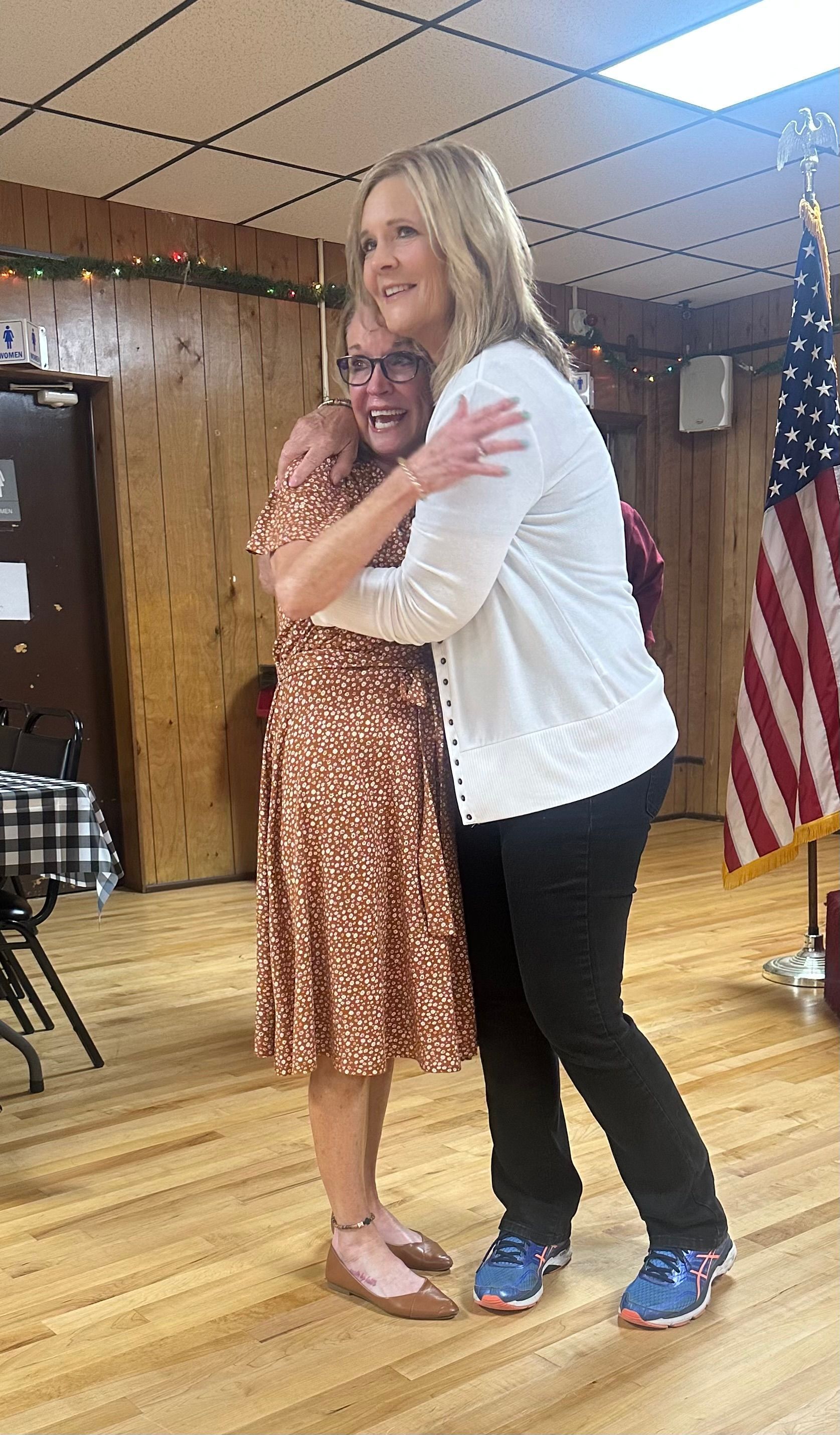 Two women are hugging each other in a room with an american flag in the background.