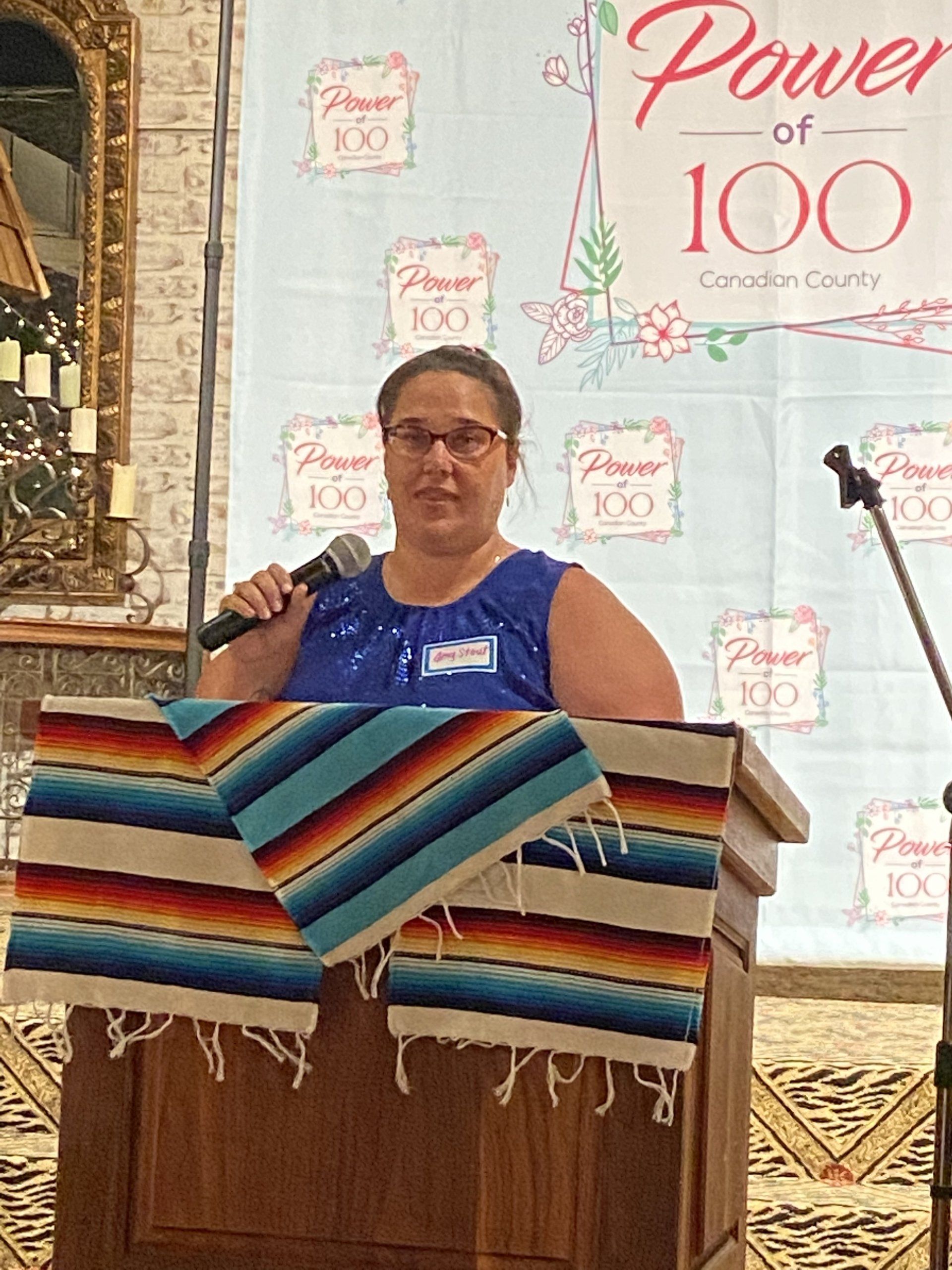 A woman stands at a podium holding a microphone in front of a sign that says power of 100