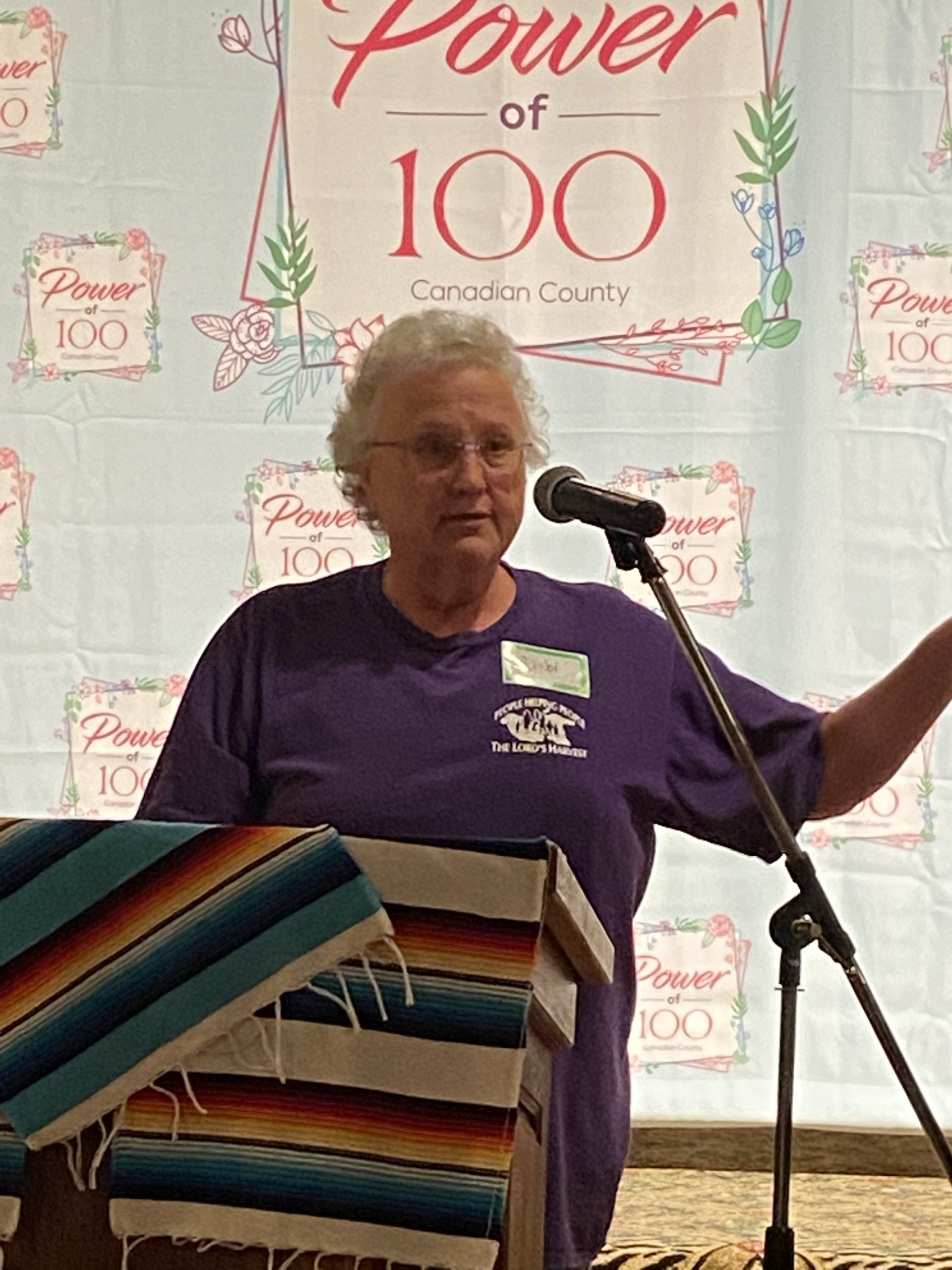 A woman in a purple shirt is speaking into a microphone in front of a sign that says power of 100