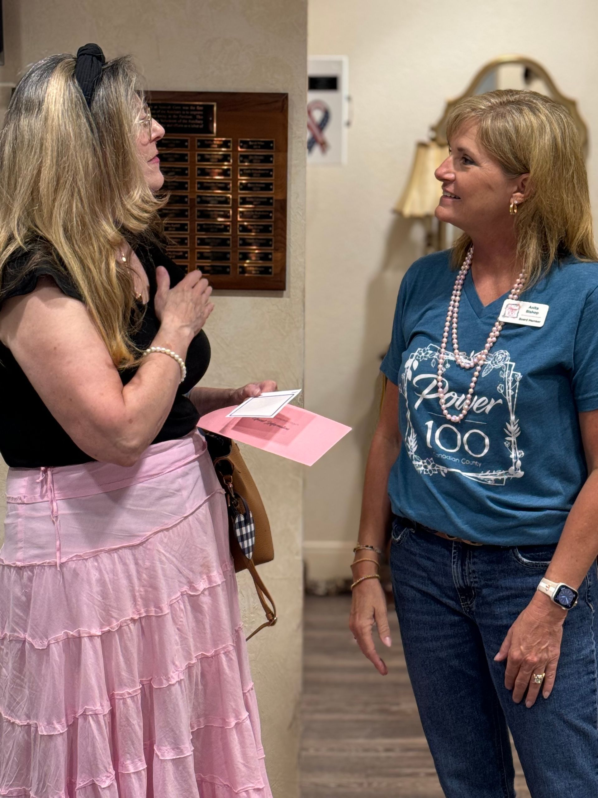 Two women are standing next to each other in a room talking to each other.