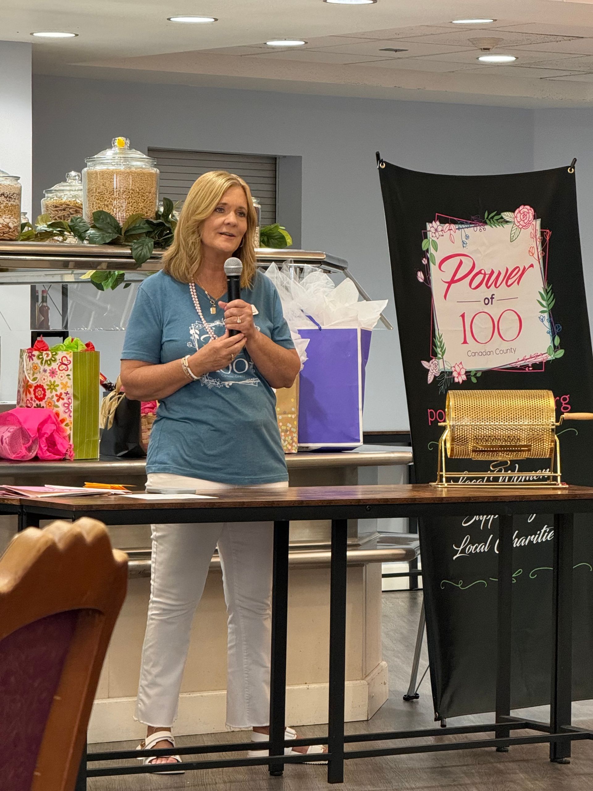 A woman is standing at a table with a microphone in front of a sign that says power 100