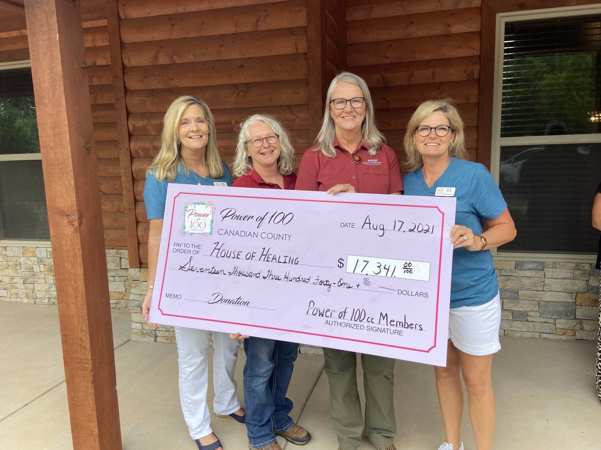 A group of women are holding a large check in front of a log cabin.