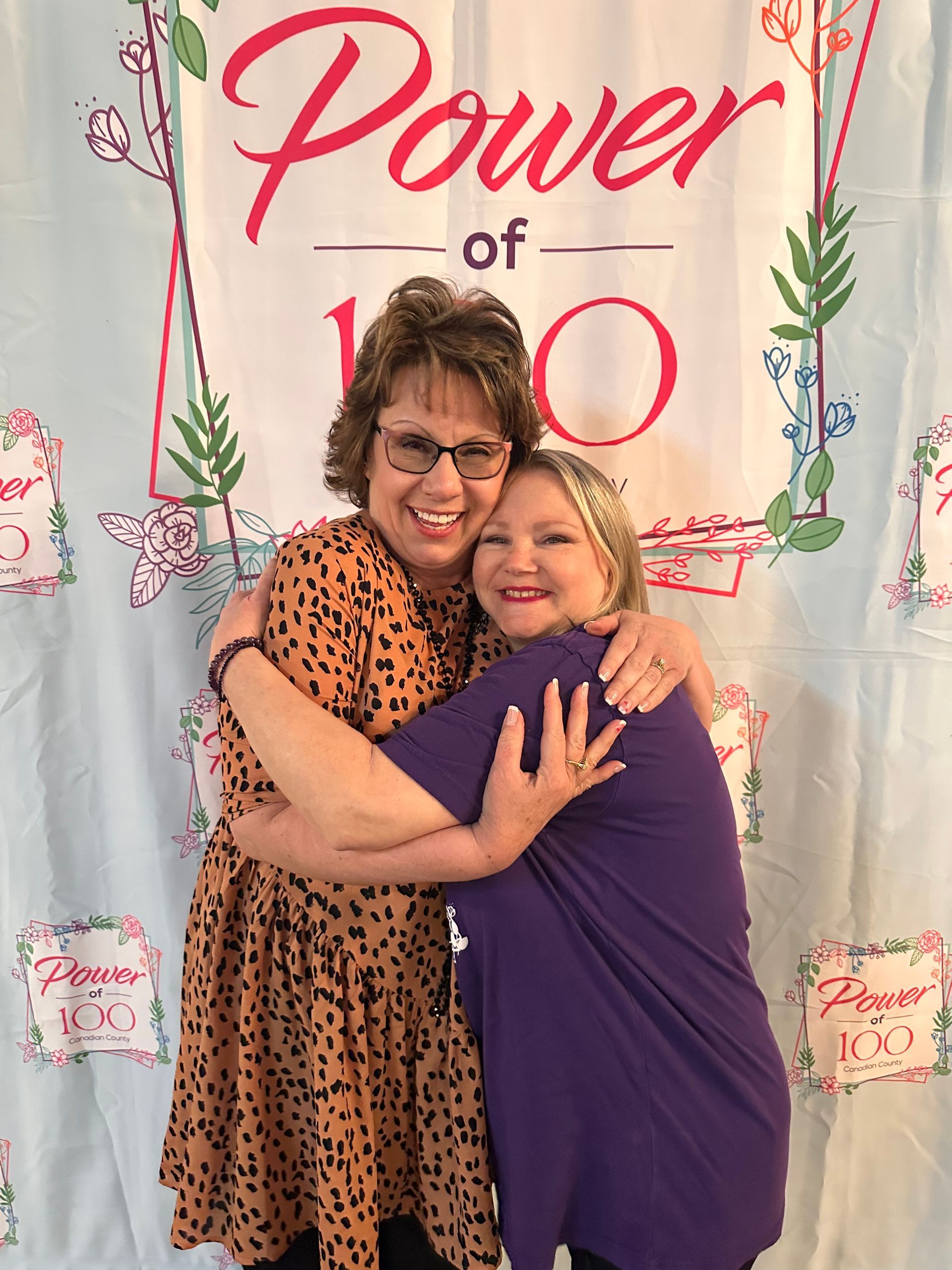 Two women hugging in front of a sign that says power of 100