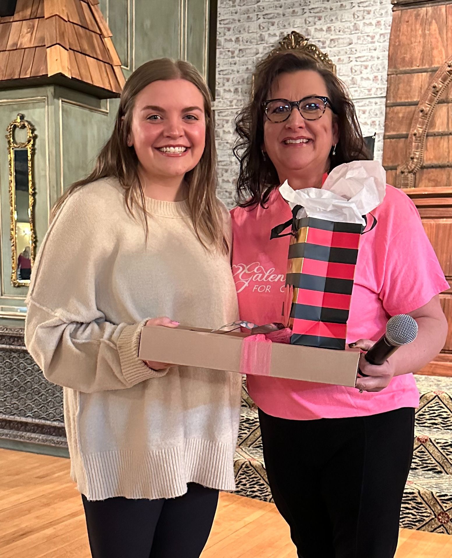 Two women are standing next to each other holding a gift box and a microphone.