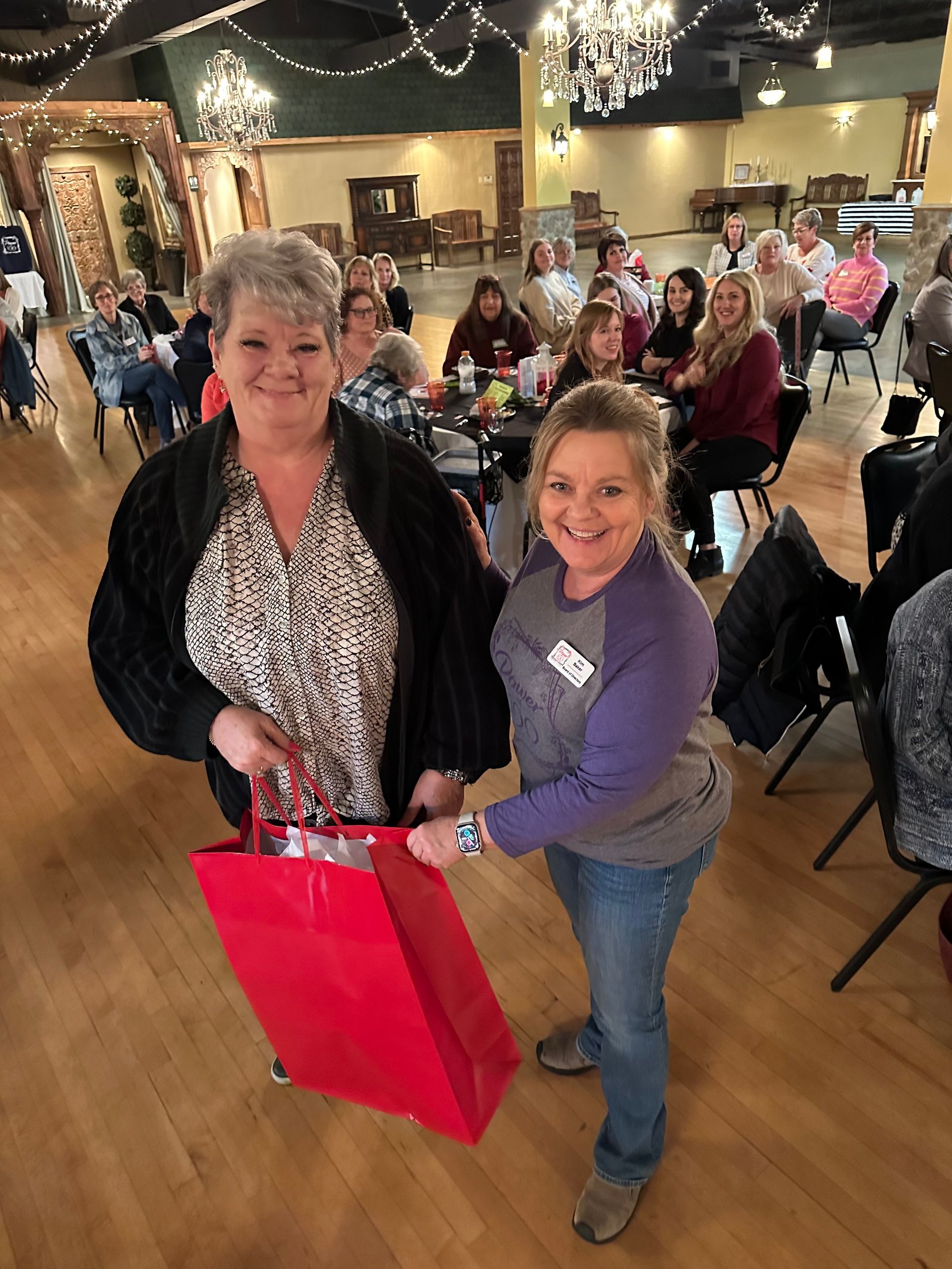 Two women are standing next to each other in a room holding red bags.