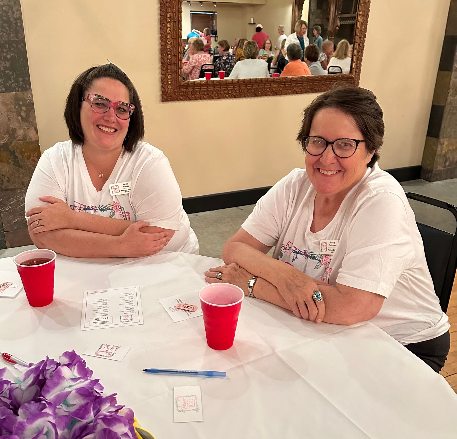 Two women are sitting at a table with purple flowers and red cups.