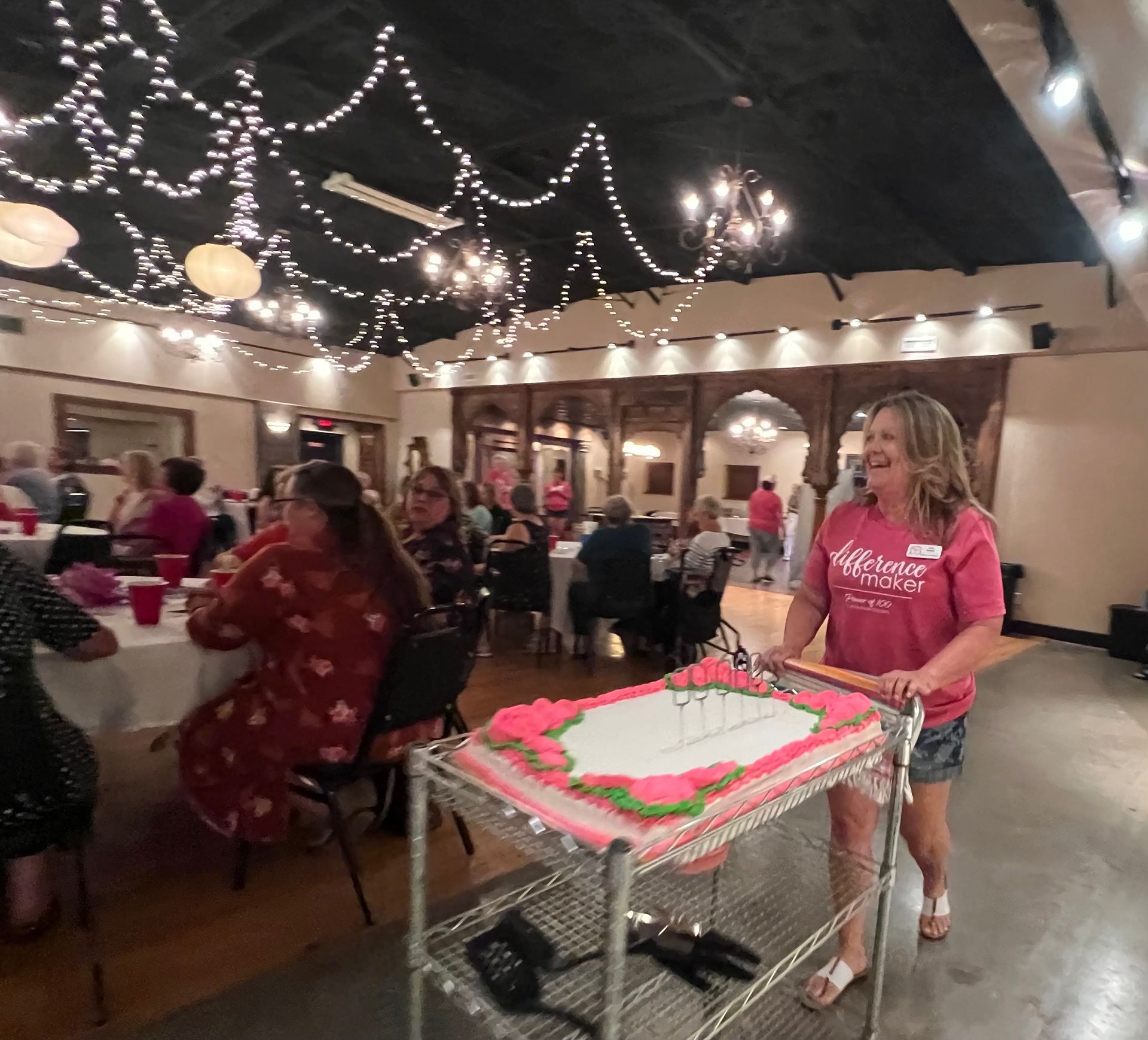 A woman is carrying a cake on a cart in a room with people sitting at tables.