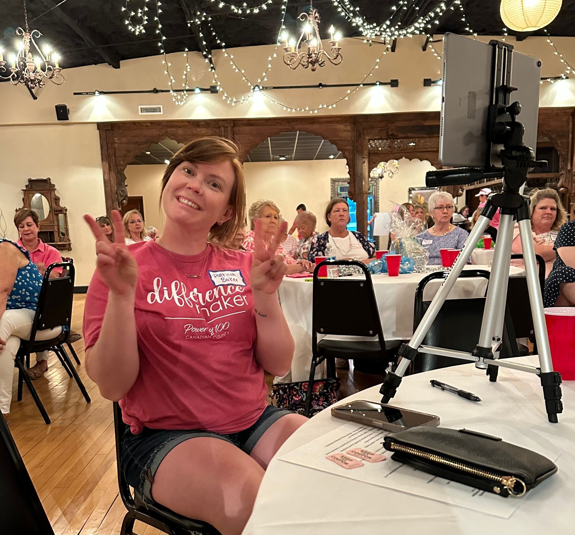A woman in a pink shirt is sitting at a table giving a peace sign.