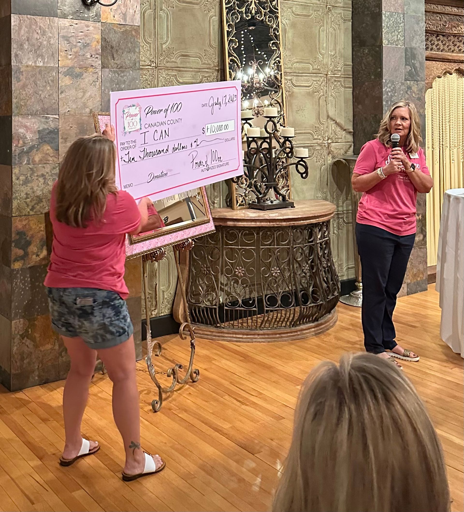 A woman in a pink shirt is holding a pink check while another woman stands behind her holding a microphone.