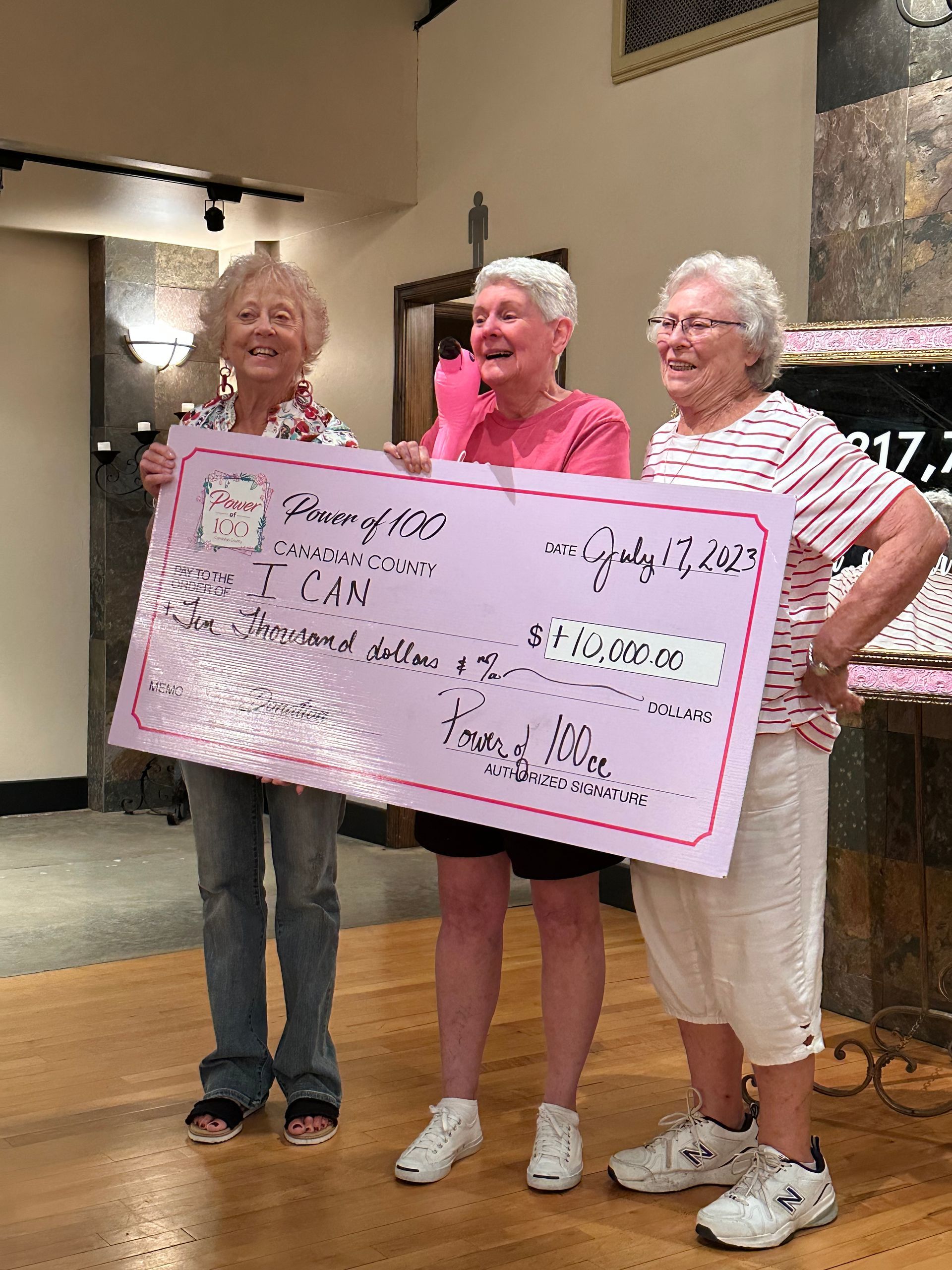 Three older women are holding a large pink check.