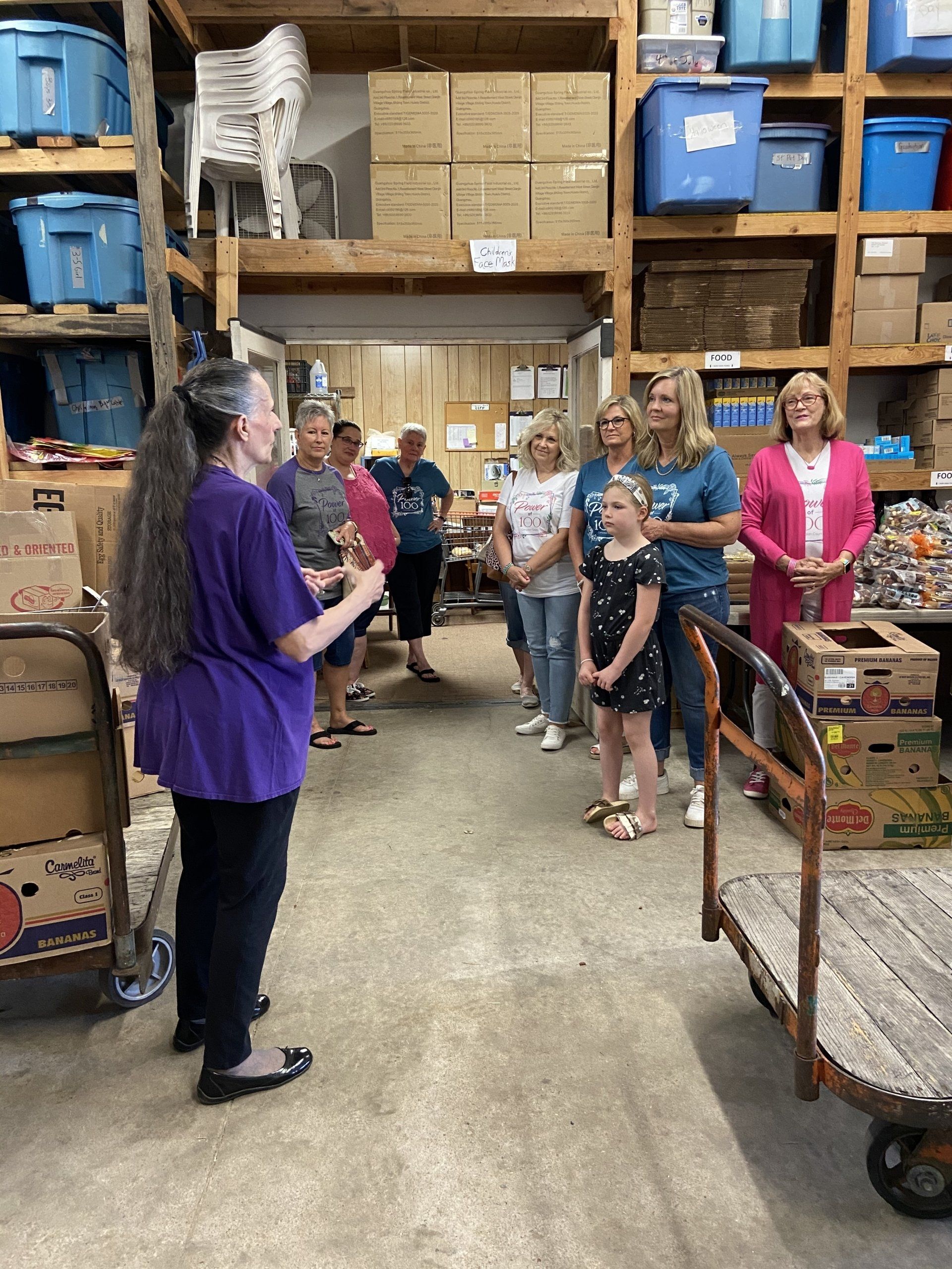 A woman in a purple shirt is talking to a group of people in a warehouse.