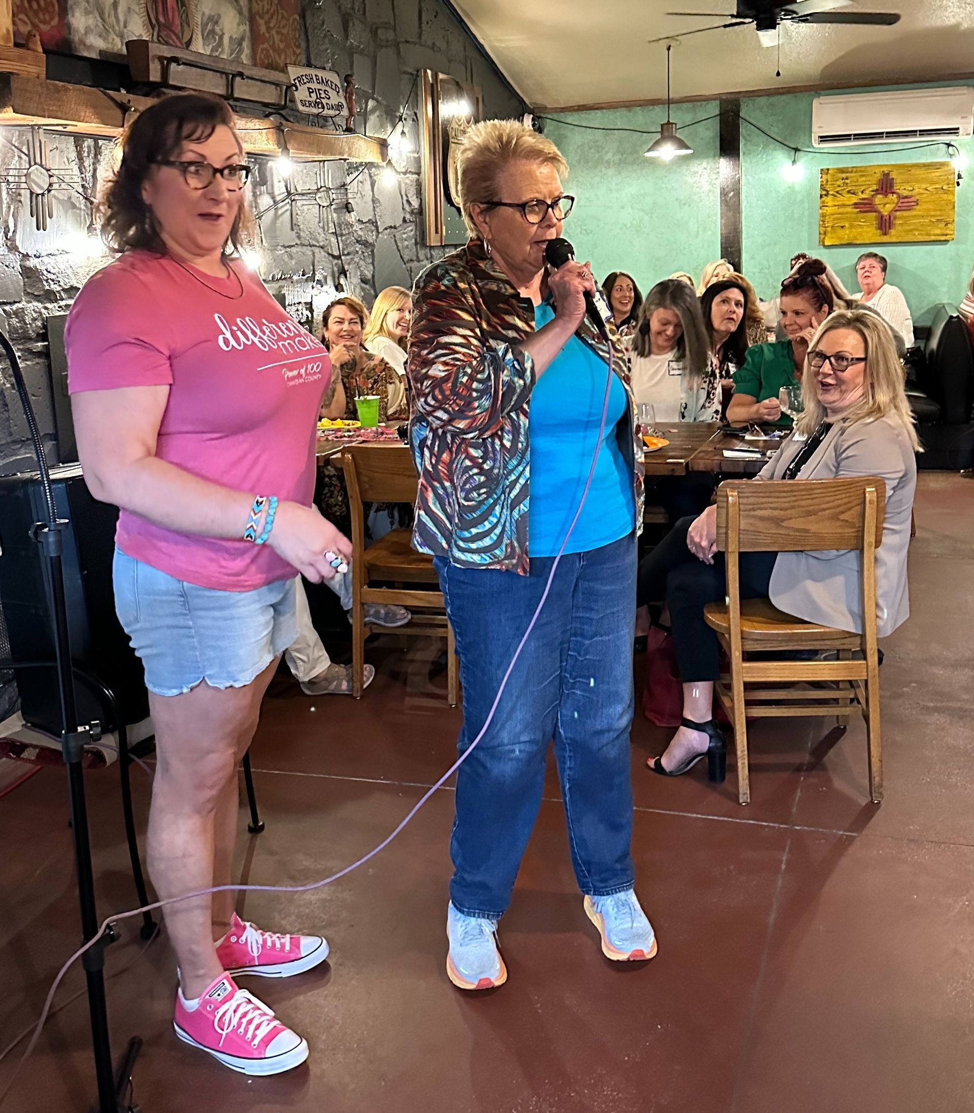 Two women are singing into microphones in front of a crowd of people in a restaurant.