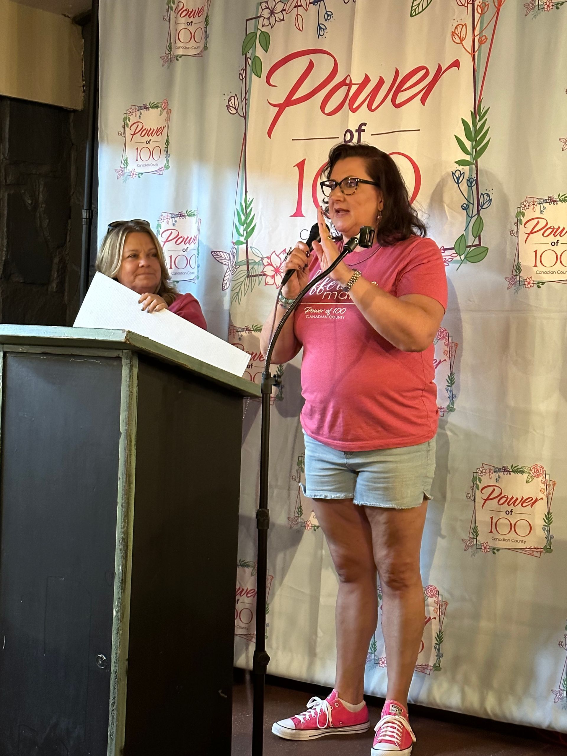 A woman in a pink shirt is standing at a podium speaking into a microphone.