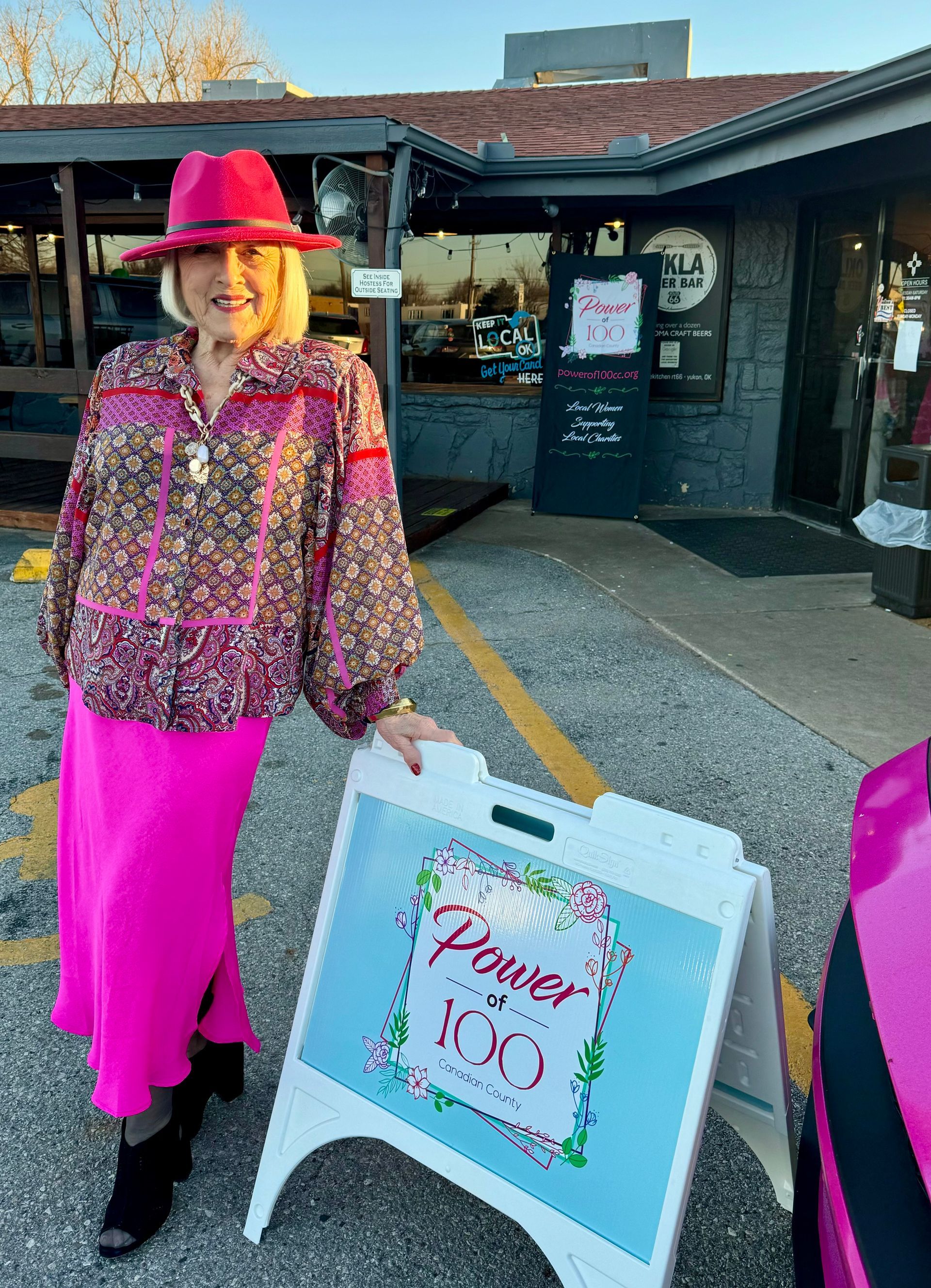 A woman in a pink skirt and red hat is standing next to a sign.