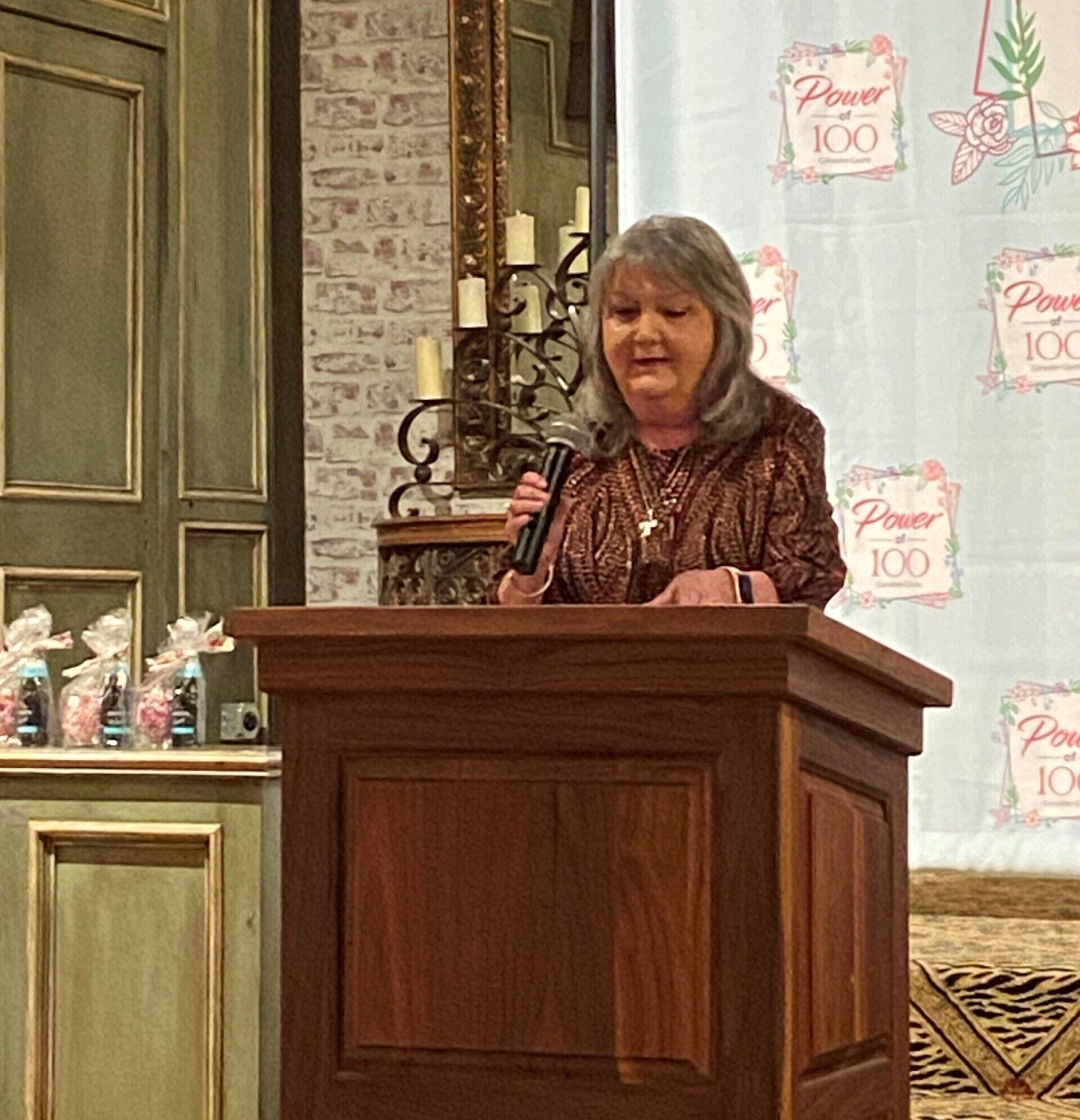 A woman stands at a podium with a microphone in front of a banner that says power 100