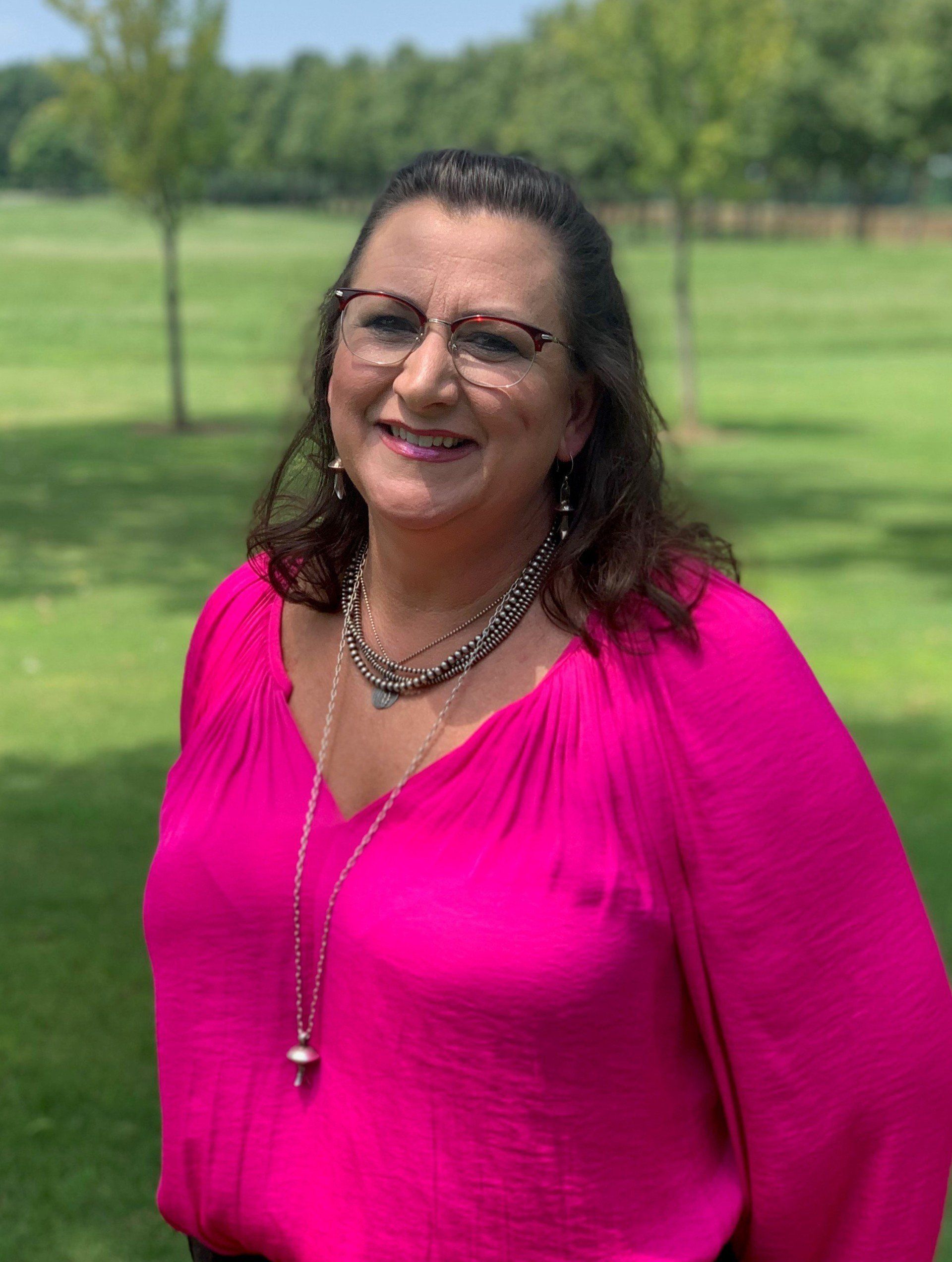A woman wearing glasses and a pink shirt is standing in a park.