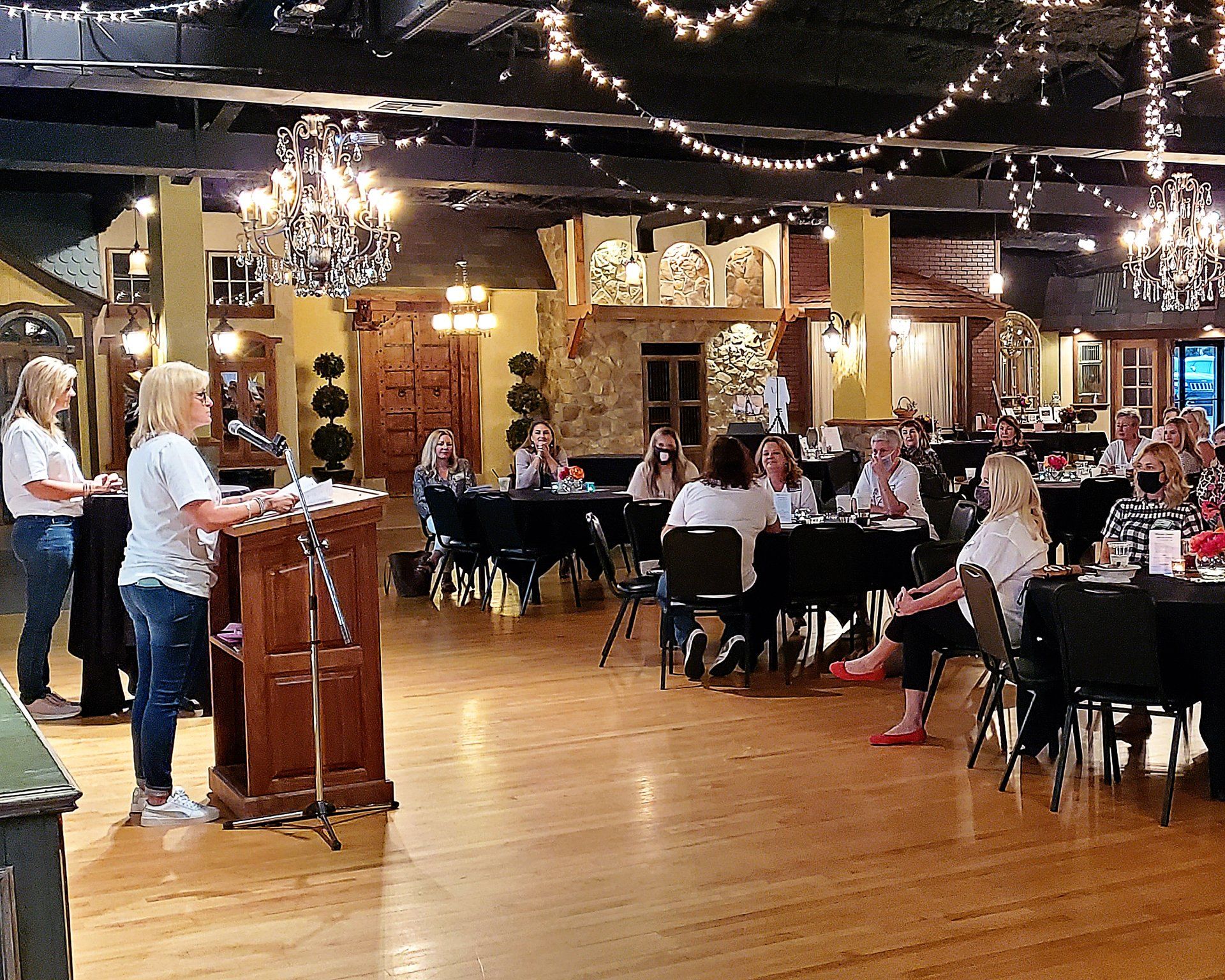 A woman is giving a speech at a podium in a large room with people sitting at tables