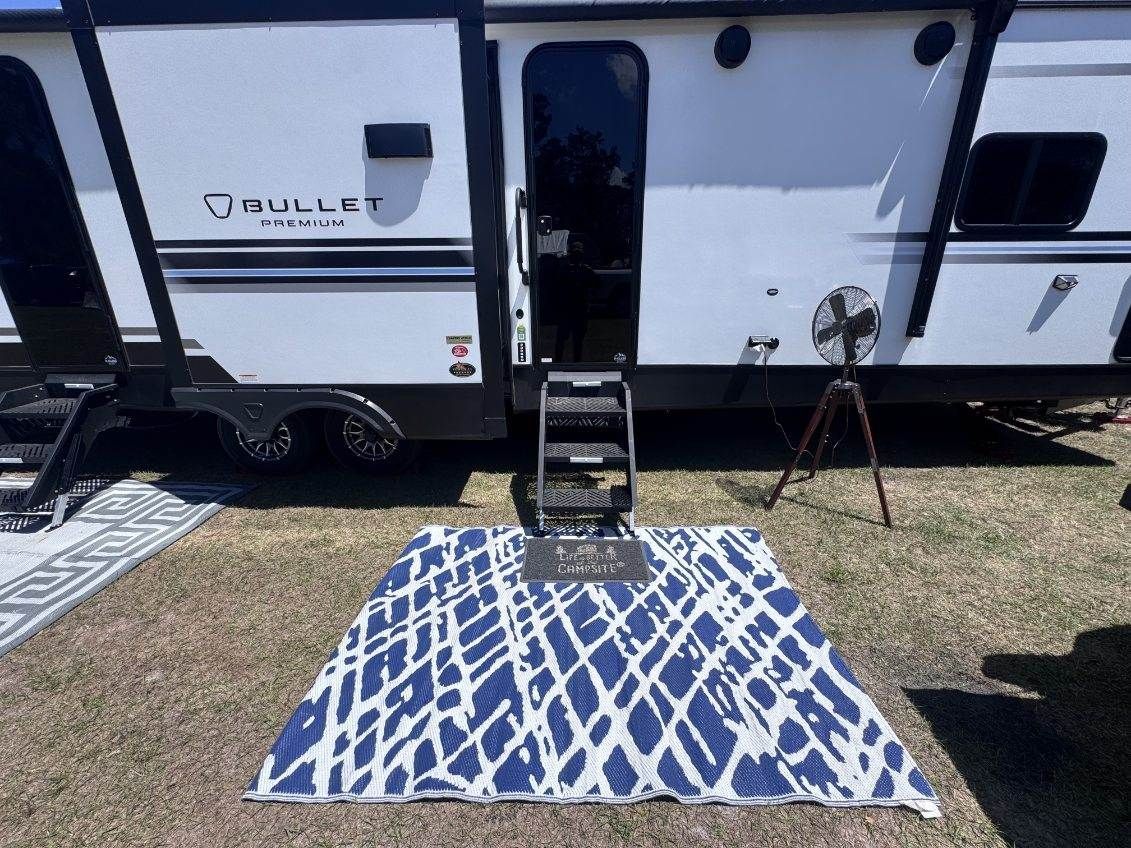 RV campsite with a blue-and-white rug, folding chair, grill, and tripod on a grassy area