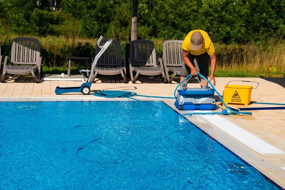 Person cleaning a blue swimming pool with a robotic cleaner, near lounge chairs and a yellow bucket.