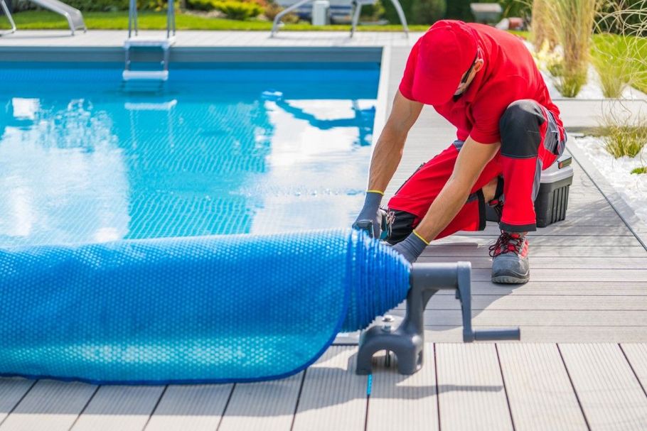 Person in red suit rolling a blue pool cover near a swimming pool.