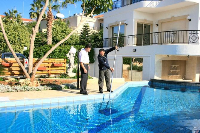 Two men cleaning a pool with a long-handled net in front of a white house.