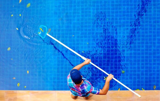 Person using a pool net to remove leaves from a blue tiled swimming pool.