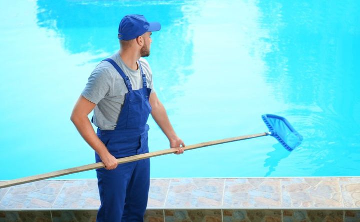 Man in blue overalls and cap using a net to clean a blue swimming pool.