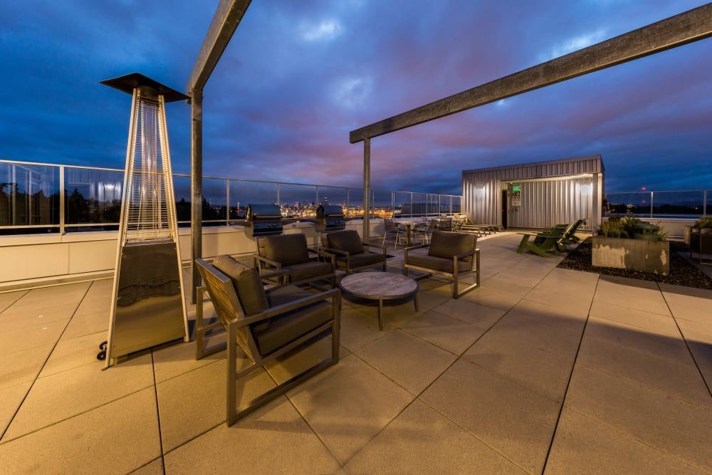 Twilight view of a modern rooftop terrace with sleek outdoor furniture including armchairs, a sofa, and coffee tables. A triangular gas fire pit adds a cozy touch. The space is complemented by minimalistic planters and a clear glass railing providing an unobstructed view of the evening skyline. Architectural beams overhead suggest a feeling of open yet intimate space at Marq West Seattle in Seattle, WA.