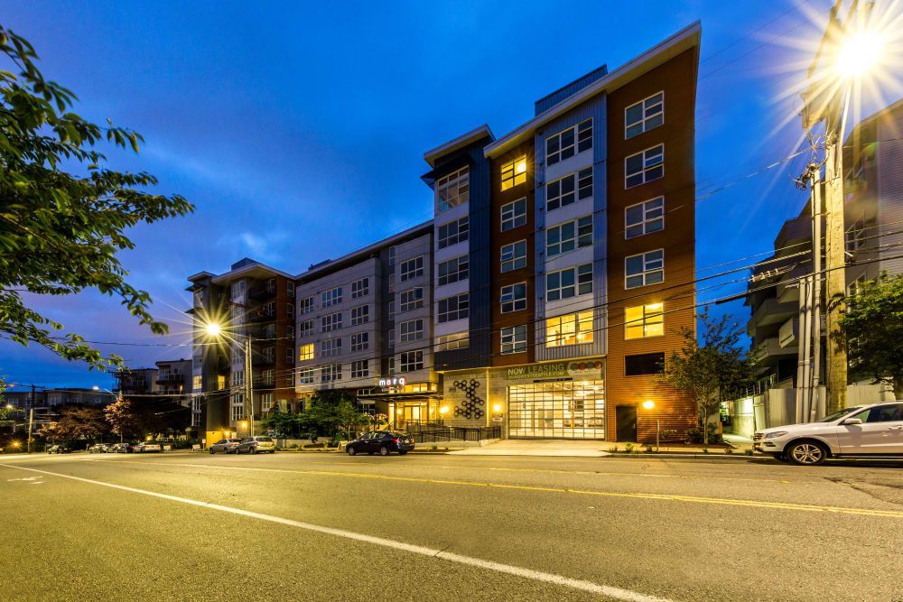 Twilight view of a modern multi-story apartment building with lit windows and a Now Leasing sign on the ground floor. A quiet street in the foreground features parked cars and a vibrant blue sky stretches above as a streetlight illuminates the scene at Marq West Seattle in Seattle, WA.