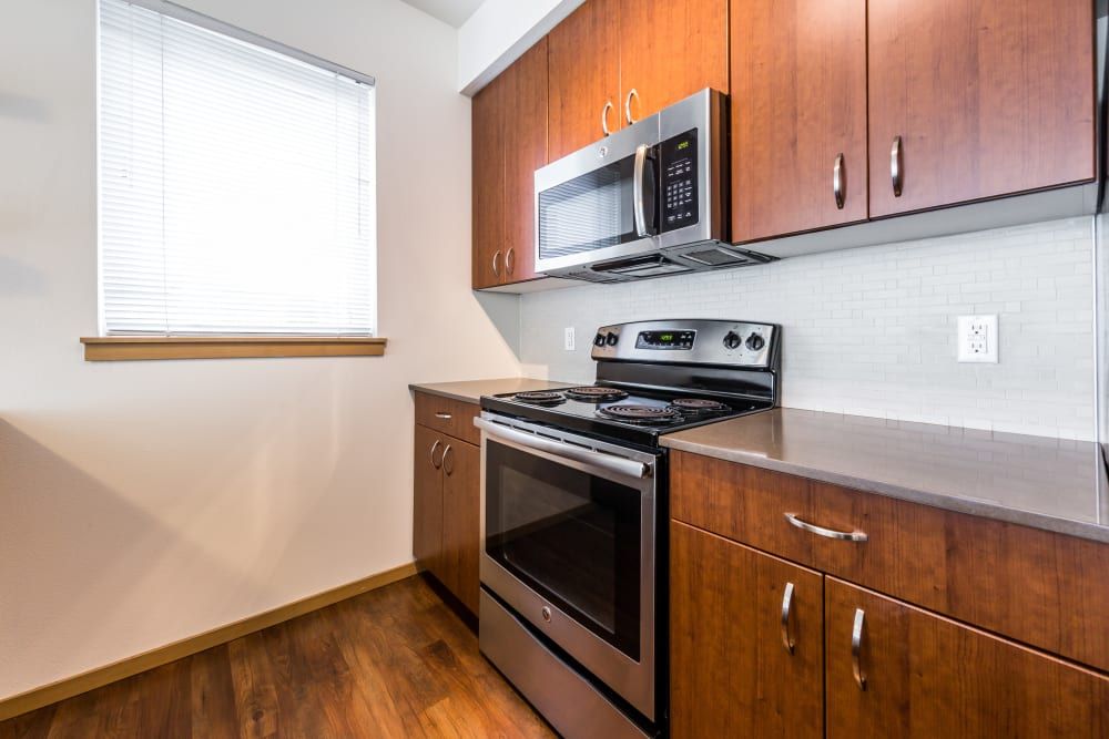 Modern kitchen interior with rich wood cabinets, stainless steel appliances including a microwave and stove, and a white subway tile backsplash. A single window with blinds allows natural light to brighten the space at Marq West Seattle in Seattle, WA.