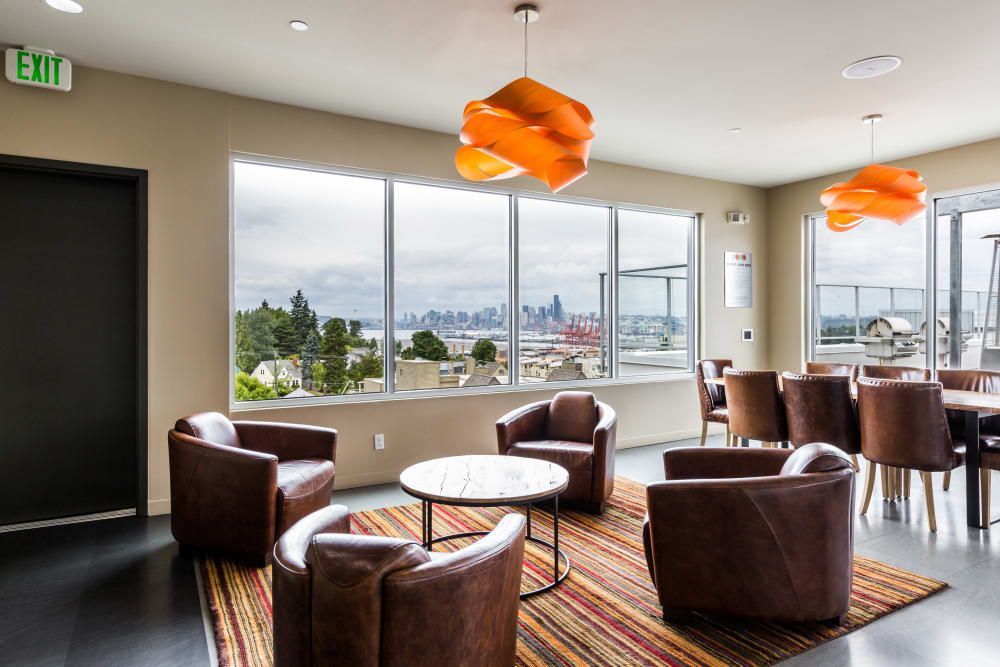 Modern room with large windows offering a view of the city skyline. The room has brown leather armchairs grouped around a white round table, an area rug with colorful stripes, and unique, orange pendant lights above. Wooden dining furniture is visible in the background, along with an EXIT sign above a doorway at Marq West Seattle in Seattle, WA.