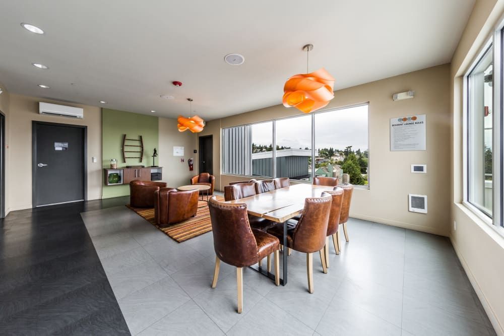 A modern and bright communal lounge area with a dining section featuring a large table and leather chairs. The room includes a sitting area with plush leather armchairs, a small coffee table, and decorative pendant lights. Large windows allow ample natural light to fill the space. The interior design is a mix of warm and neutral tones, with black flooring and green accent walls at Marq West Seattle in Seattle, WA.