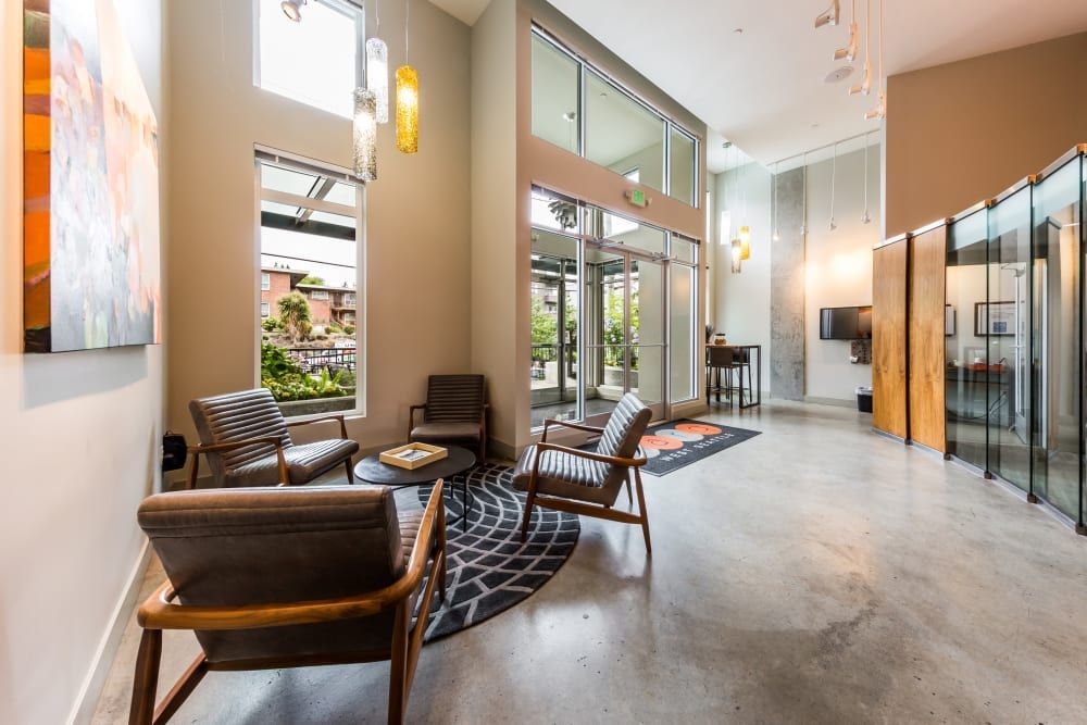 Modern waiting area with polished concrete floors. It features comfortable leather armchairs, a geometric-patterned area rug, several hanging pendant lights, and large windows providing natural light. The space has an open feel with a visible outdoor courtyard, glass partition doors, and a reception desk in the background at Marq West Seattle in Seattle, WA.