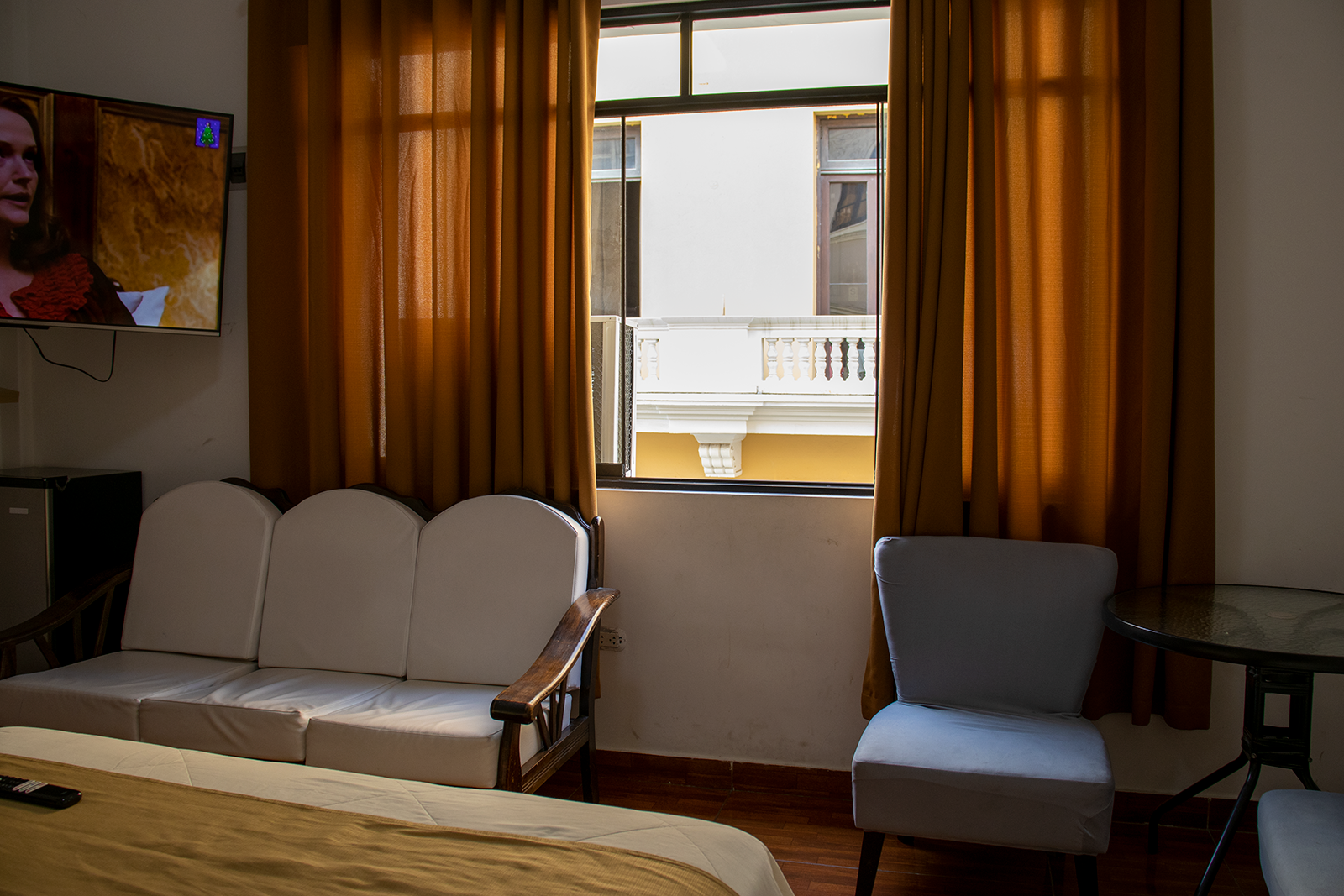 Interior de habitación de hotel: sofá blanco, silla gris, ventana con cortinas, TV.