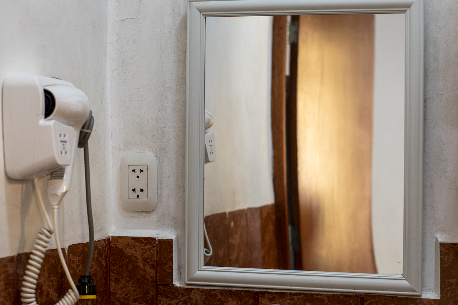 Secador de pelo blanco y enchufe en la pared, junto a un espejo. Zócalo de azulejos marrones.