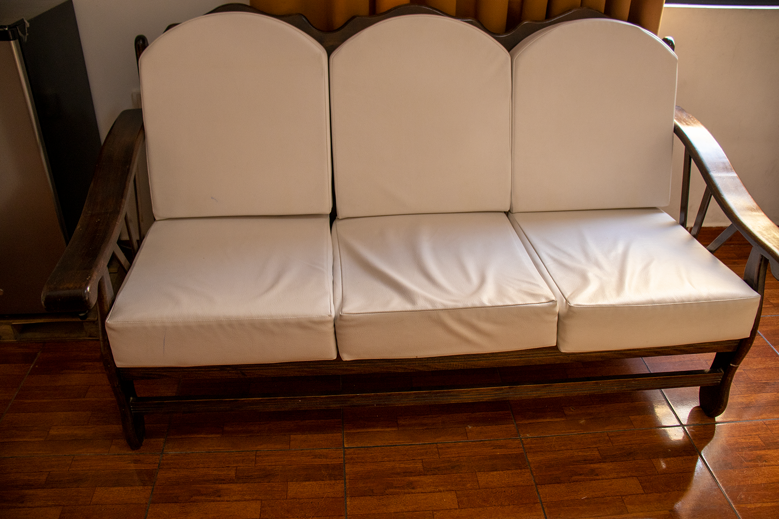 Un sofá de tres plazas de madera con cojines blancos sobre un suelo de baldosas marrones.