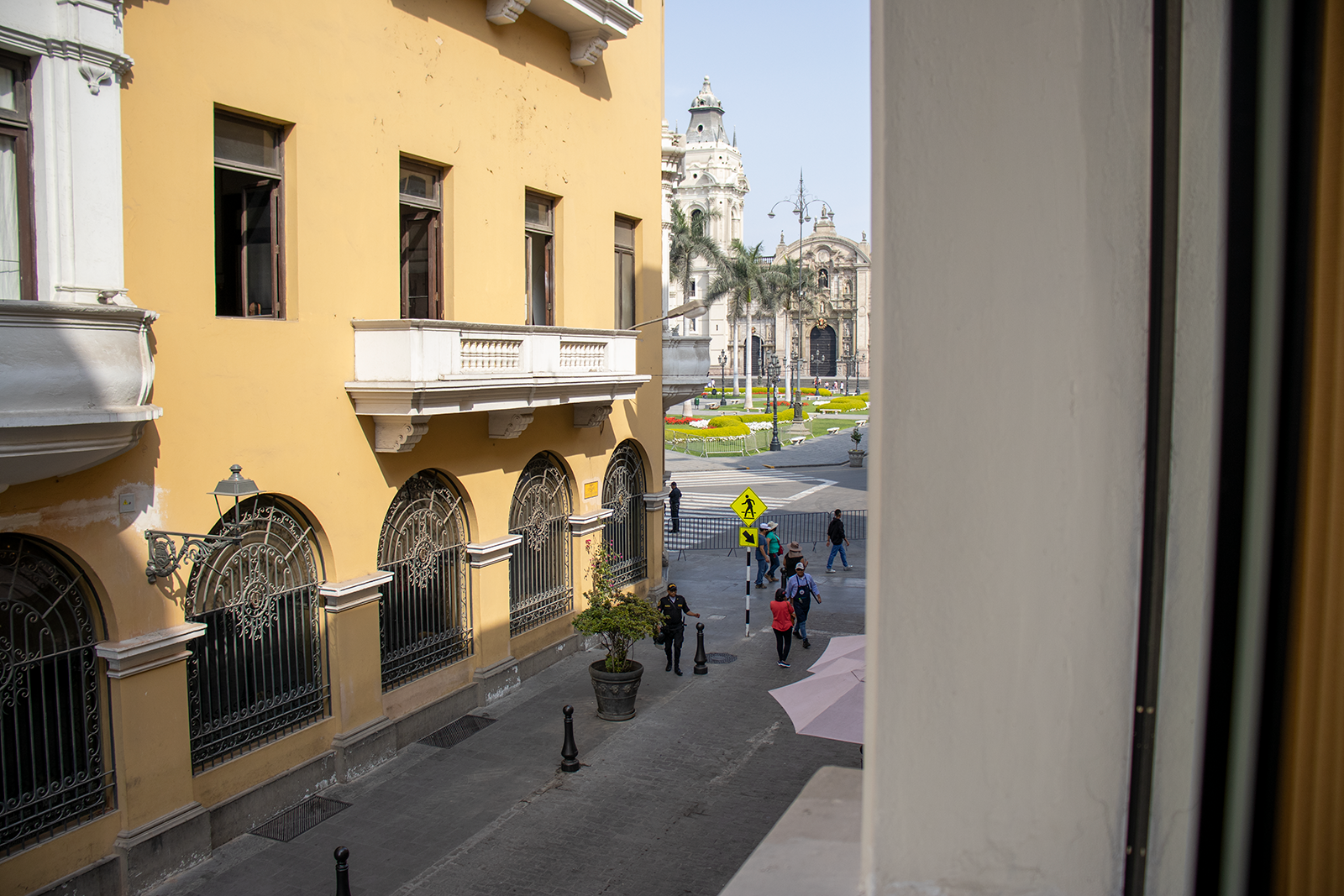 Edificio amarillo con ventanas de hierro frente a calle y plaza con gente; iglesia blanca al fondo.
