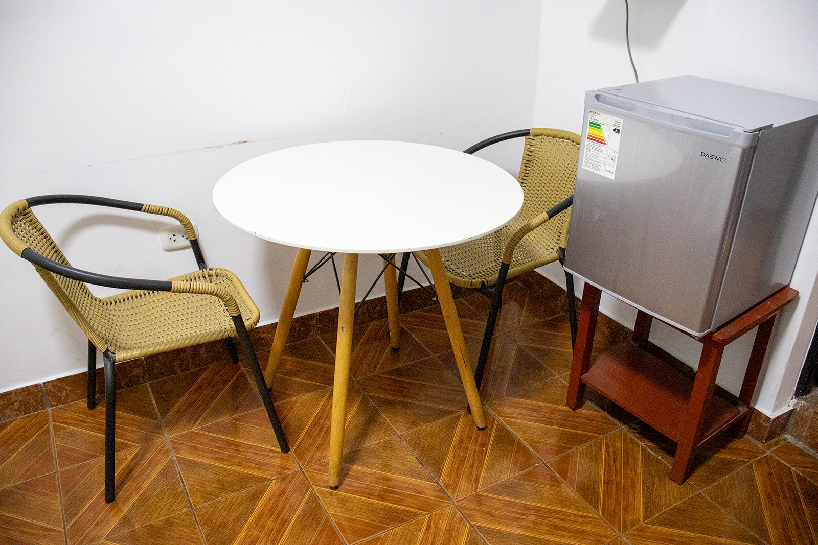 Pequeña mesa redonda con dos sillas y una mininevera en una habitación con suelo de baldosas.