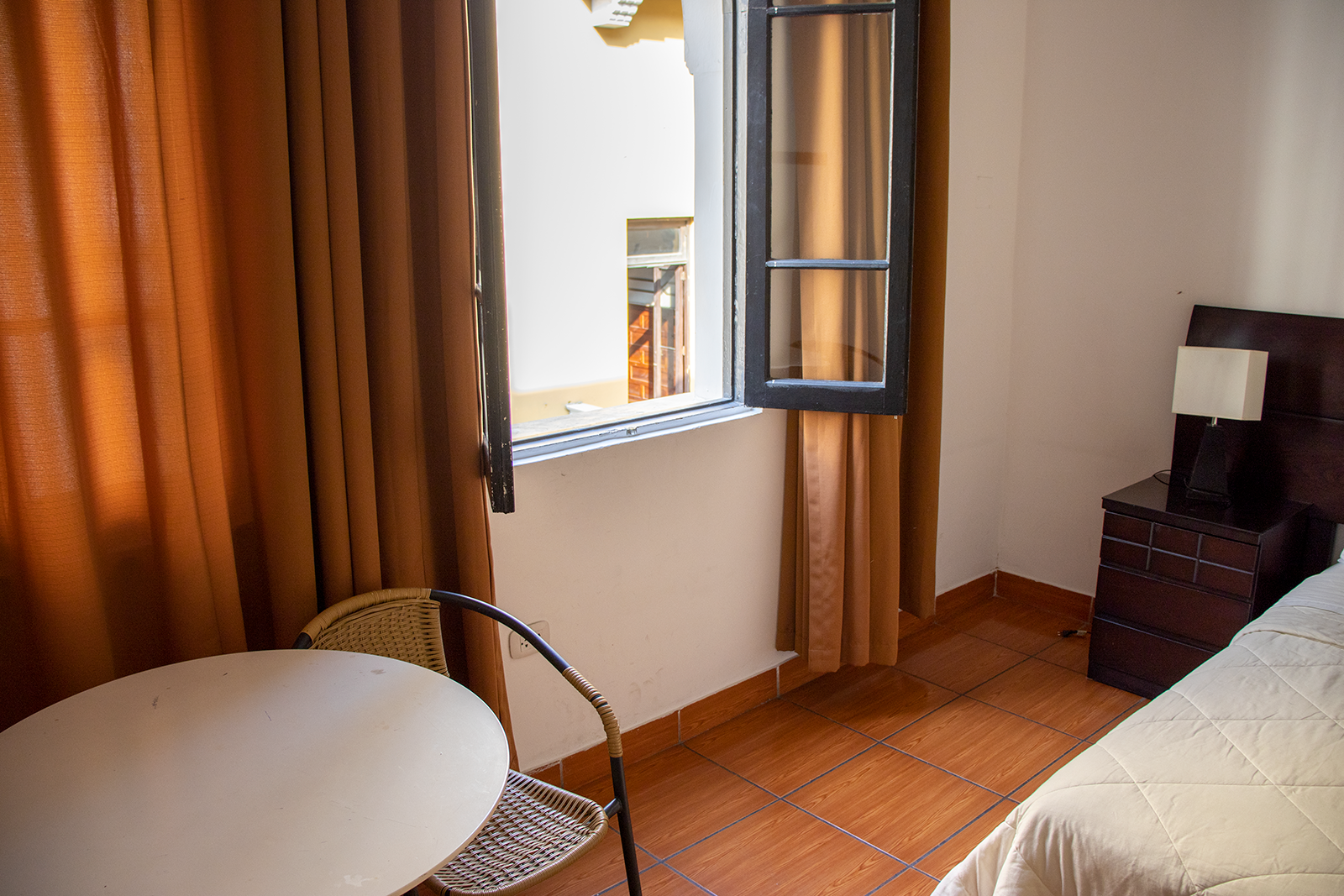 Pequeña habitación de hotel con ventana y cortinas. Mesa, silla y mesita de noche con lámpara.