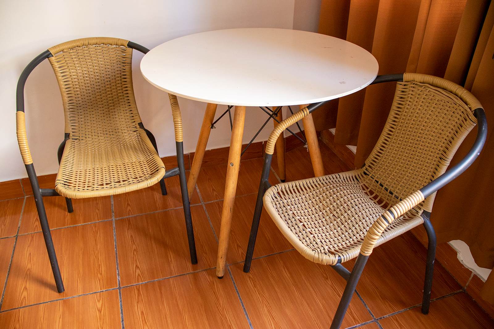 Dos sillas de mimbre frente a una pequeña mesa blanca con patas de madera, en una habitación con azulejos marrones.